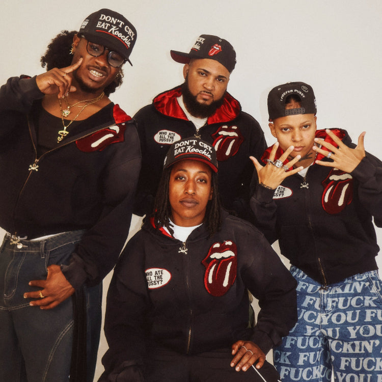 Four people posing together in matching black Rolling Stones-themed zip-up tracksuits with the iconic red tongue logo on the chest, black caps, and one person wearing a blue patterned skirt.