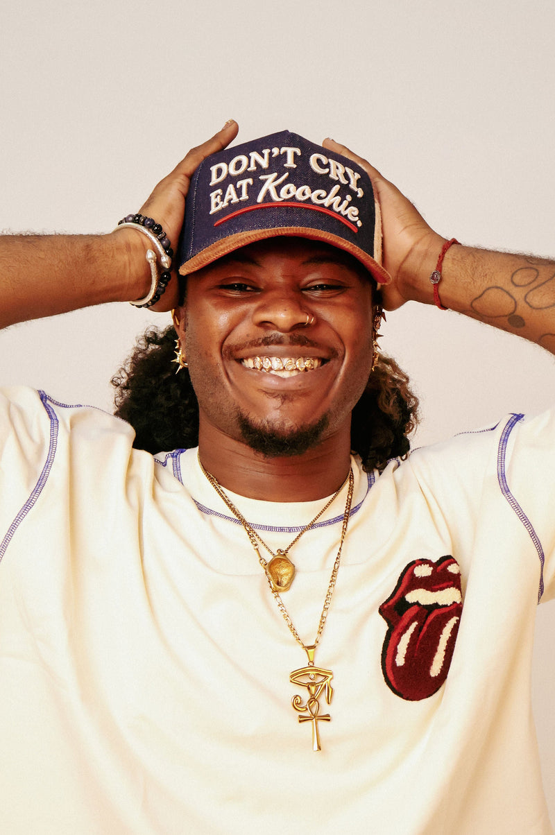 A smiling man wearing a navy blue baseball cap with the text 'DON'T CRY, EAT Koochie.' in white, a cream-colored Rolling Stones-style shirt with purple trim and a red tongue-and-loops logo, and gold necklaces, posing with his hands on his cap against a plain light background.